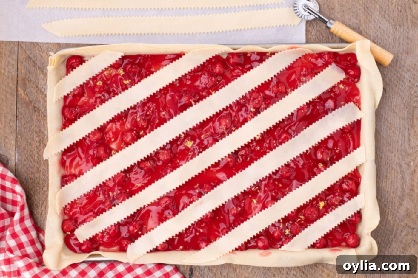 The first few strips of pie crust are carefully layered horizontally over the cherry filling in the slab pie, starting the lattice pattern.
