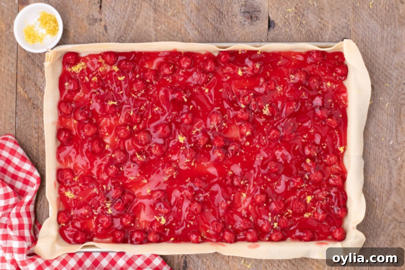 Sweet cherry pie filling spread evenly over the pie crust in a jelly roll pan, with lemon zest sprinkled on top, ready for the lattice crust.