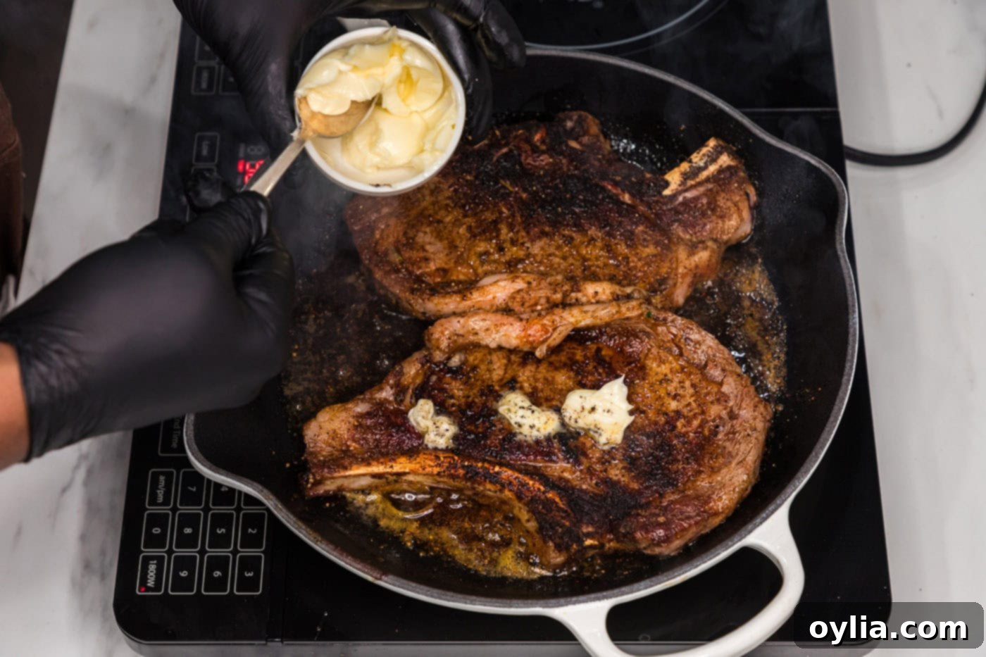 Adding pats of butter on top of sizzling bone-in ribeye steaks in a skillet for rich flavor.