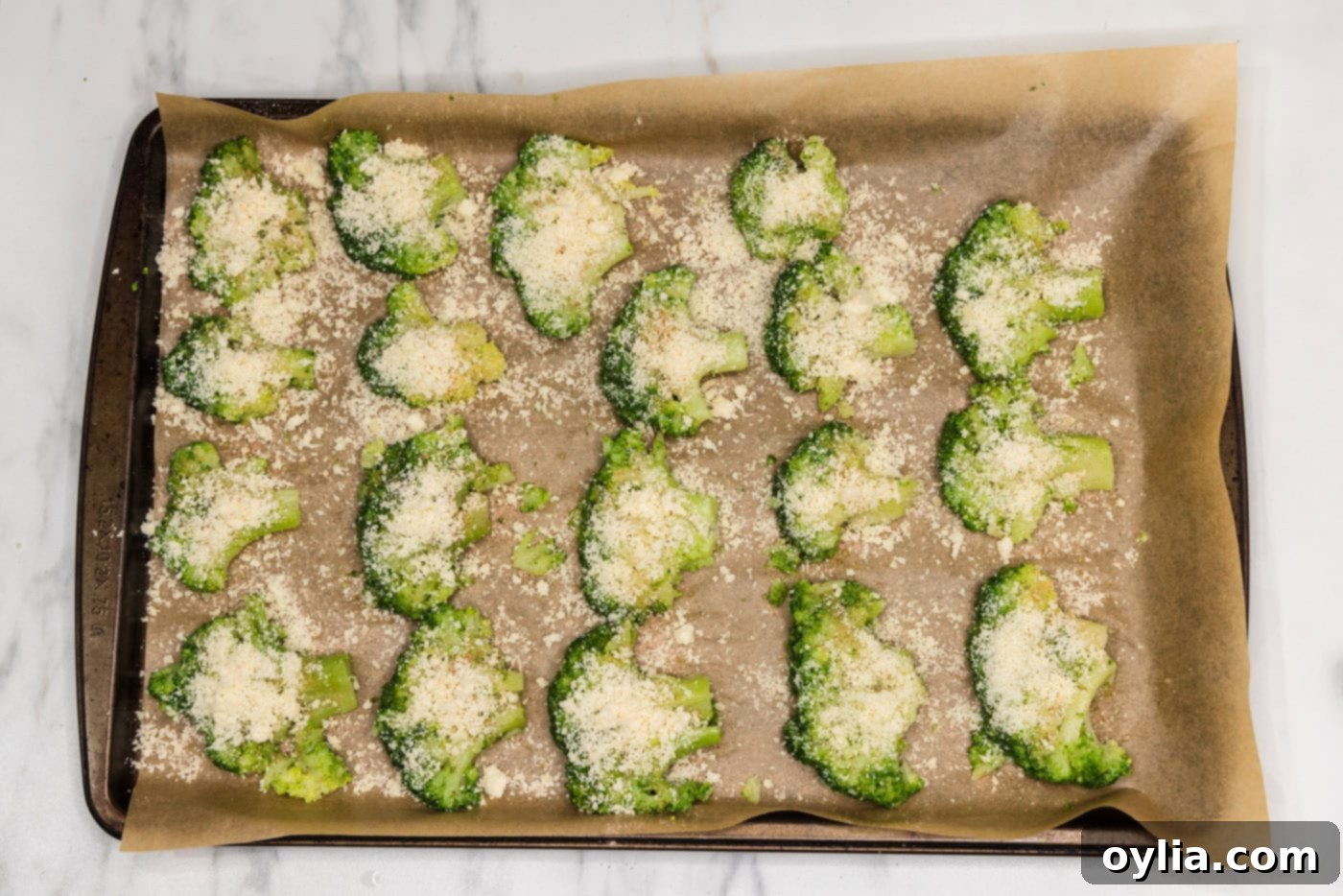 Smashed broccoli florets on a baking sheet, topped with grated parmesan cheese before baking.