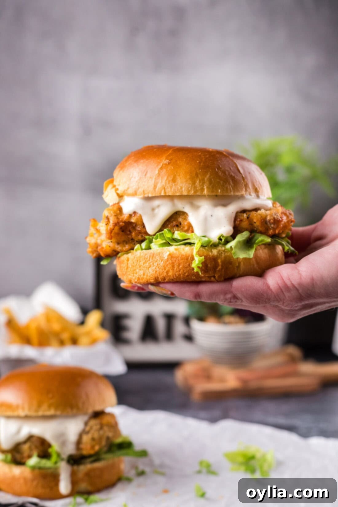 A hand holding up a freshly made Crispy Chicken Burger, showcasing the fluffy bun, crispy chicken, and vibrant toppings.