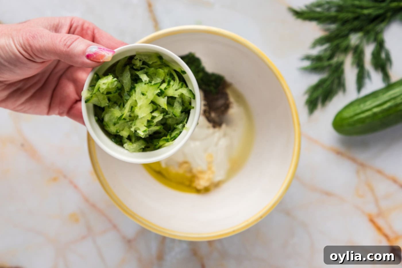 adding shredded cucumber to a bowl of tzatziki sauce ingredients
