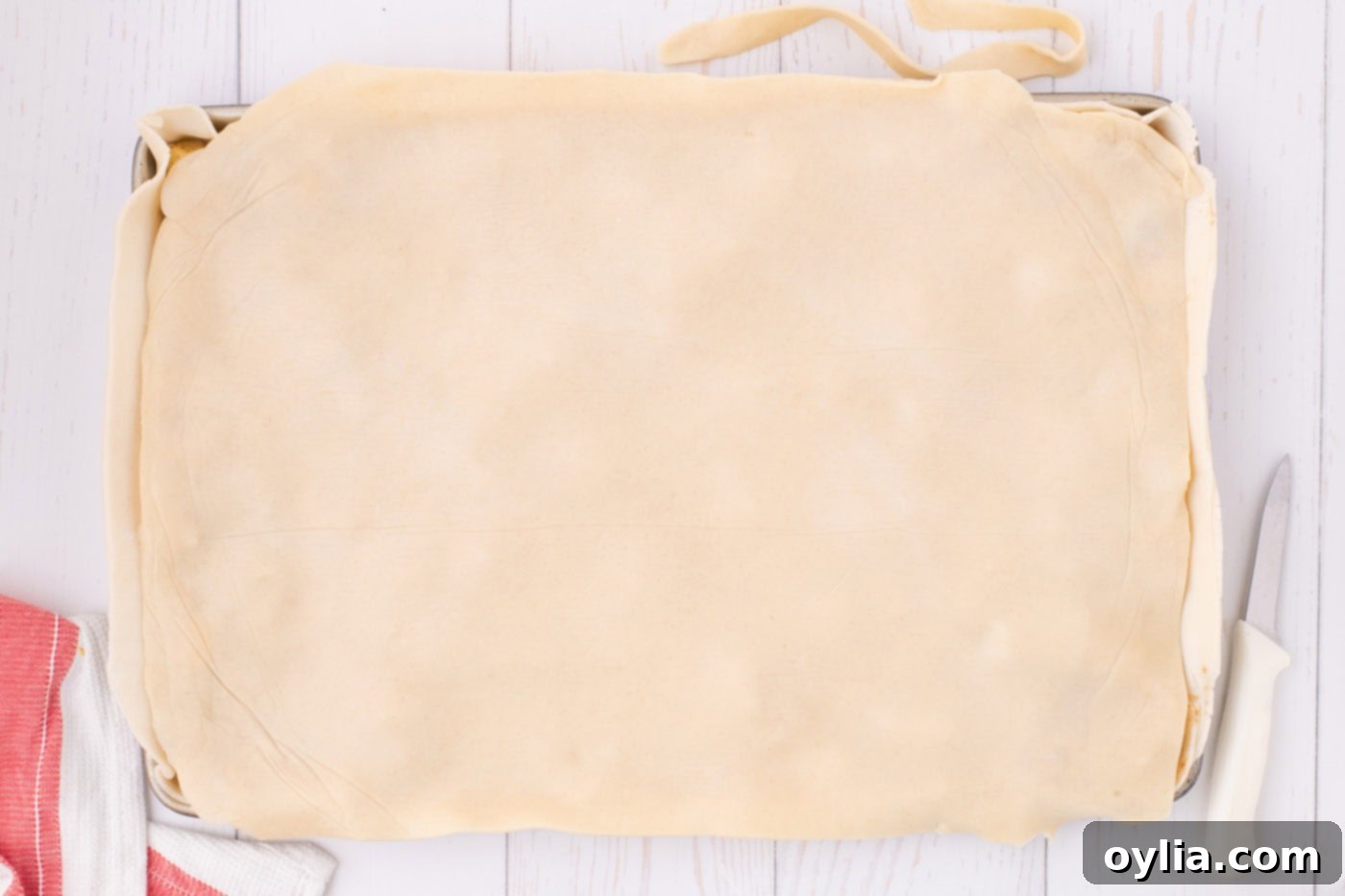 The top pie crust carefully placed over the spiced apple filling in a jelly roll pan, ready to be sealed, with excess dough trimmed.