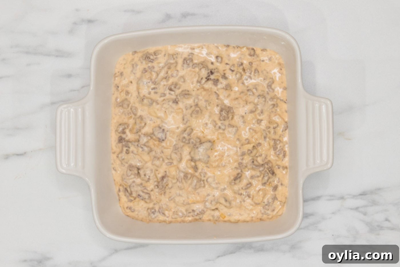 The cheeseburger dip mixture evenly spread in an 8x8 baking dish, ready for the final layer of cheese.