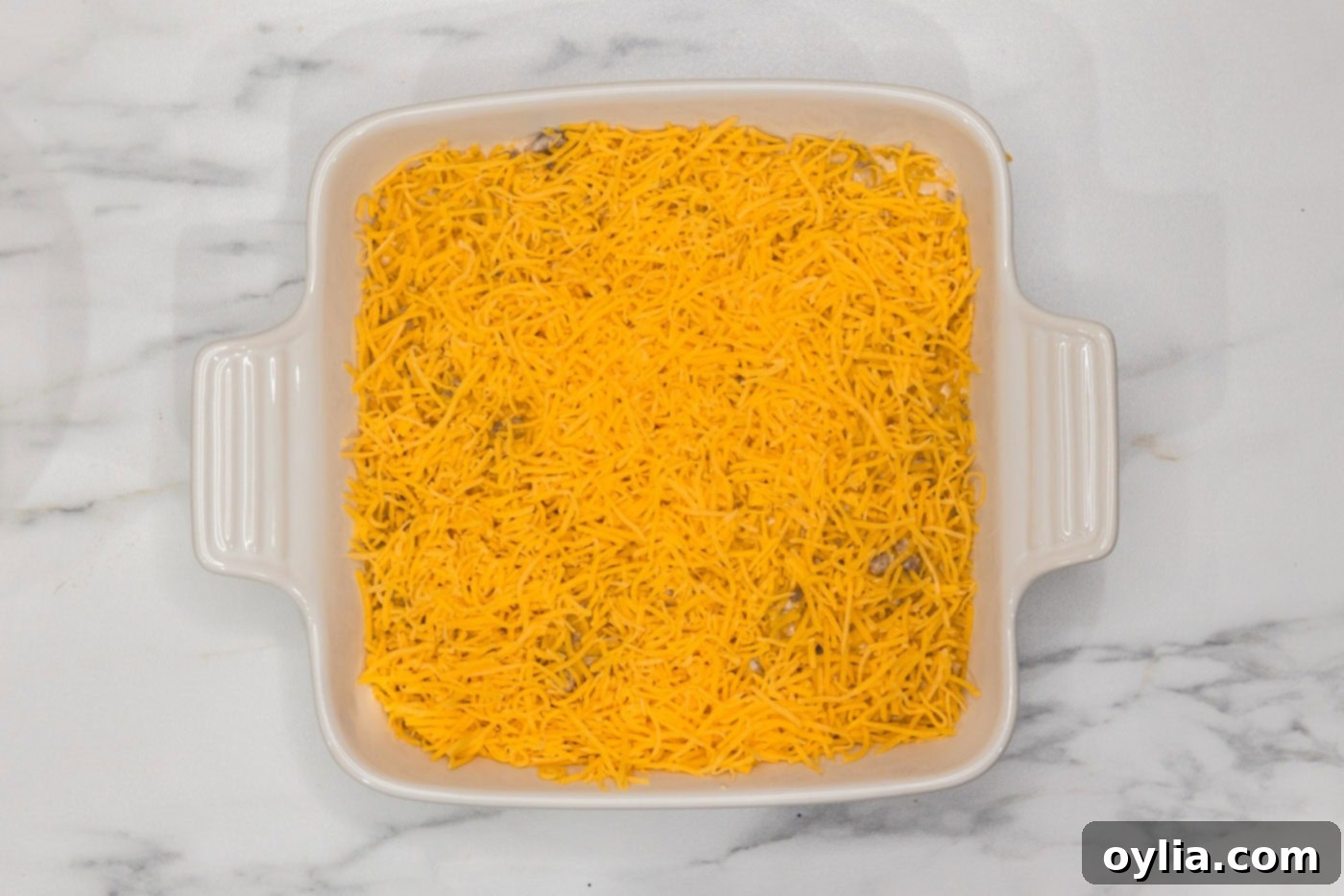 A generous layer of shredded cheddar cheese covering the cheeseburger dip in the baking dish, awaiting baking.