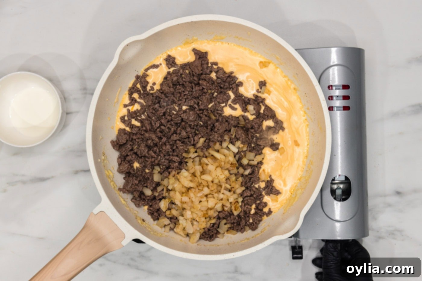 Ground beef and sautéed onions being added to the creamy cheese sauce in a skillet.