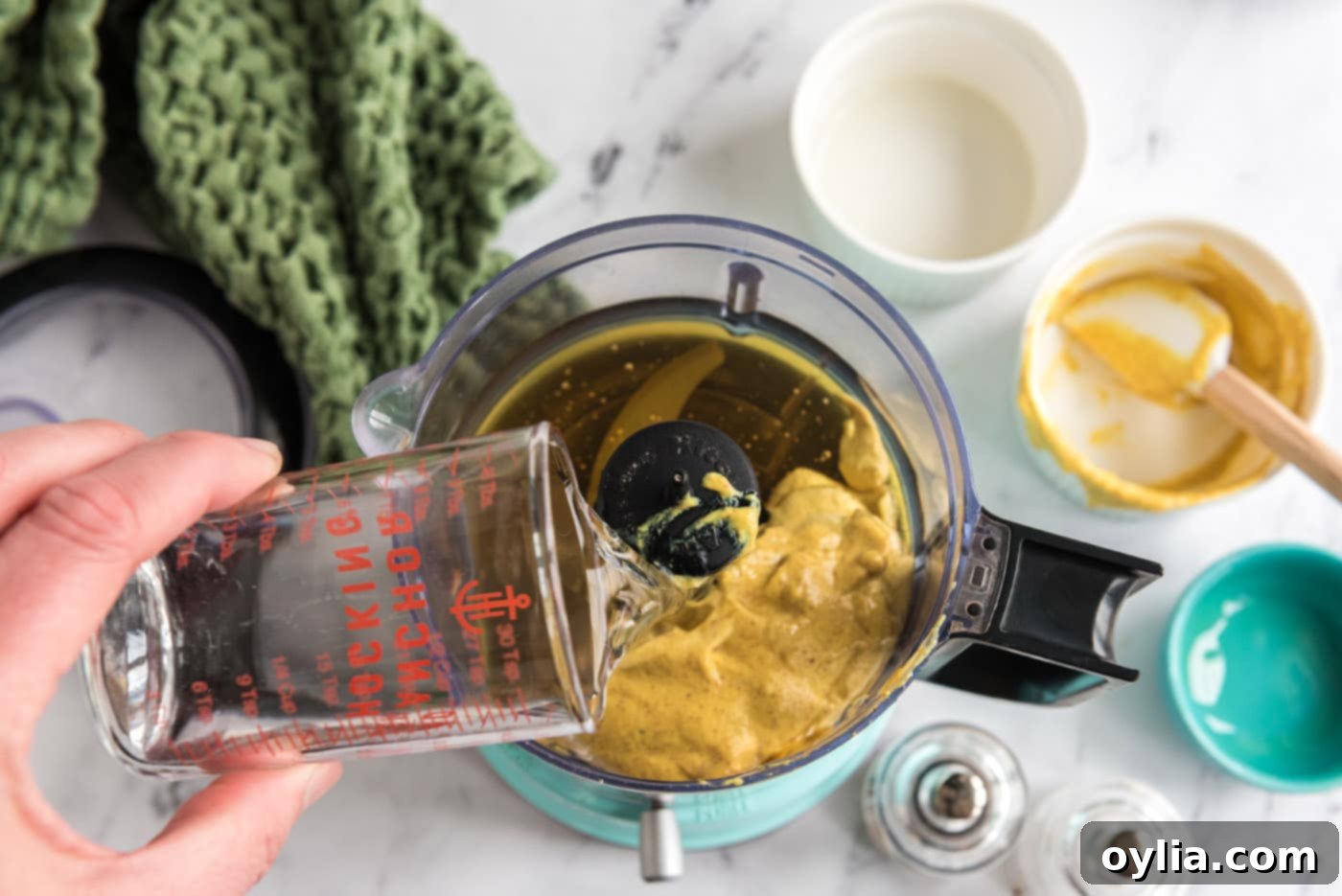pouring water into food processor with mustard, olive oil, and honey, preparing the dressing