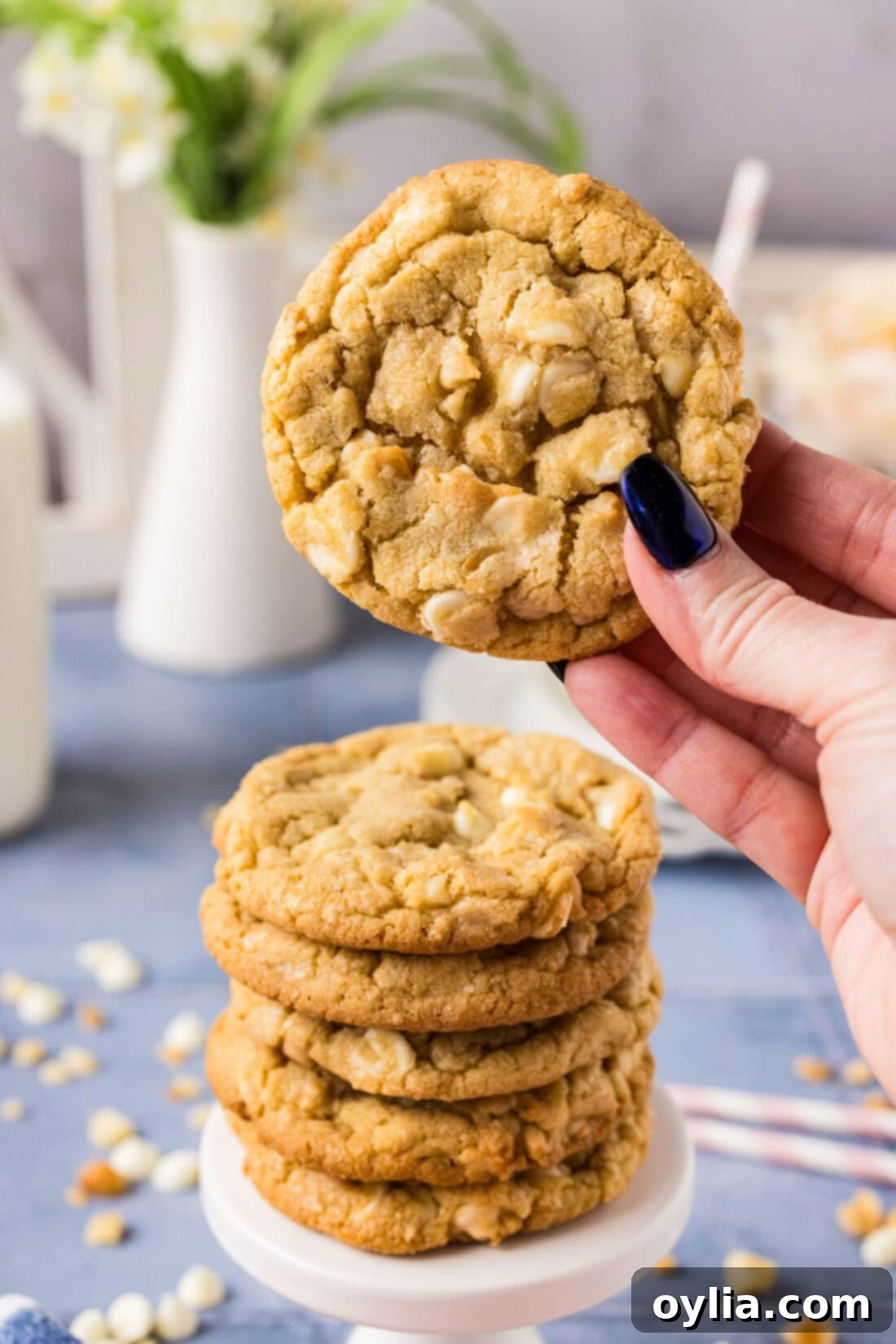 Hand holding up a White Chocolate Macadamia Nut Cookie above a stack of more cookies, showcasing its soft texture and generous fillings.