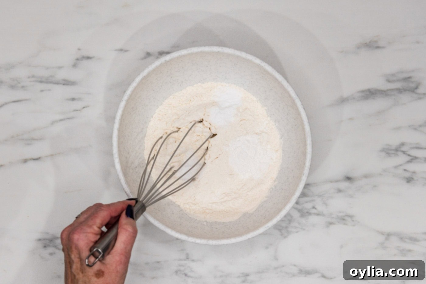 Hand whisking flour, baking powder, baking soda, and salt in a bowl, showing the dry ingredients mixing.