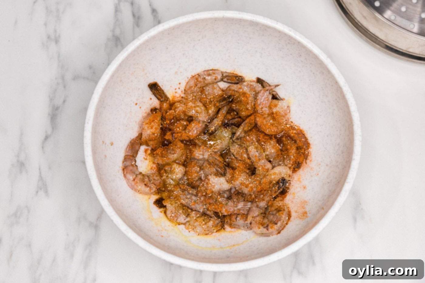 Raw shrimp in a bowl with Old Bay seasoning and olive oil, being tossed to coat