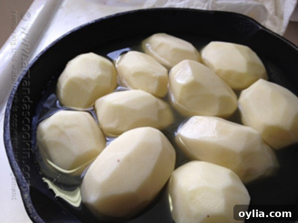 A close-up shot of carefully peeled russet potatoes nestled in a cast iron skillet, with broth added to about one inch up the sides.