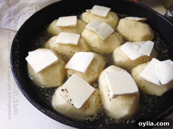 Peeled russet potatoes in a cast iron skillet, perfectly seasoned with garlic salt and pepper, topped with thin slices of cold butter, ready for roasting.