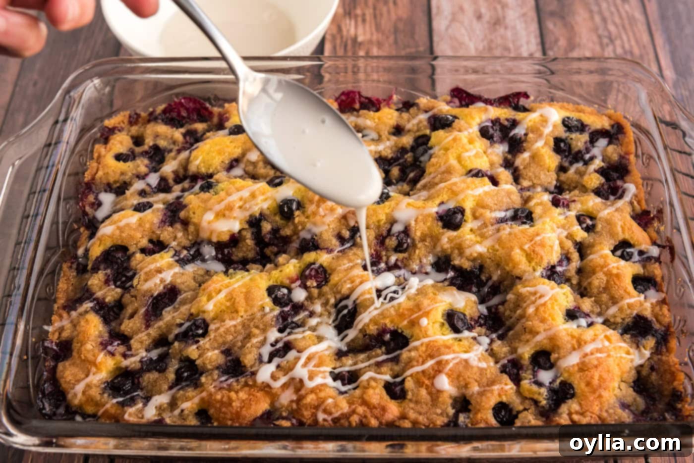 Glaze being drizzled over the cooled lemon blueberry cake, creating a beautiful finish.