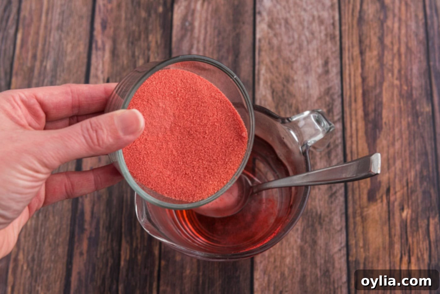 Hand pouring powdered raspberry gelatin into a bowl of boiling water, preparing the Jell-O mixture.