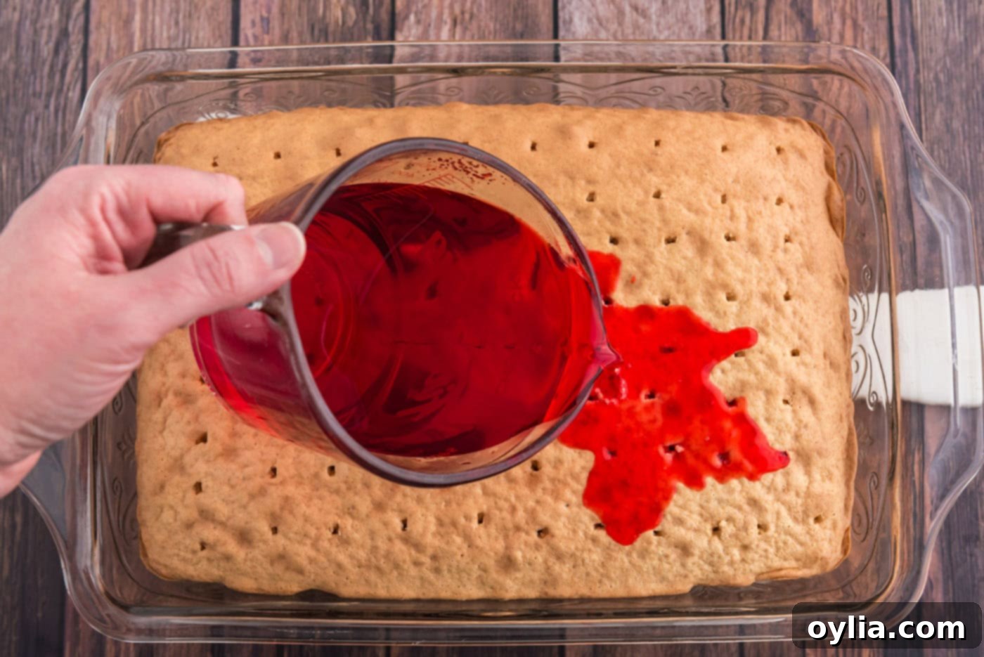 Close-up of vibrant red raspberry Jell-O being slowly poured over the poked white cake, allowing it to soak into the holes.