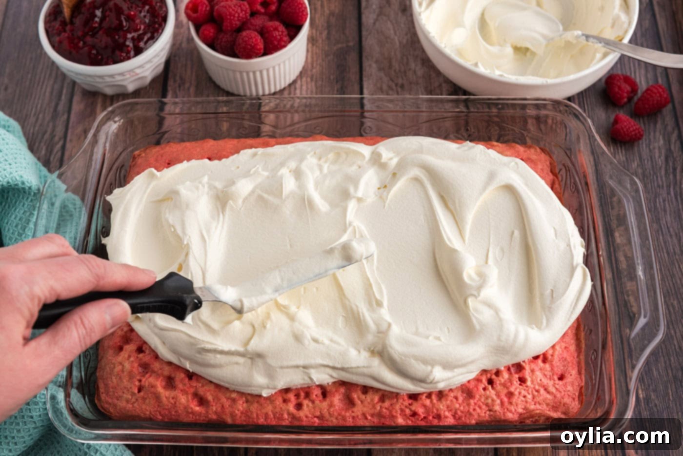 An offset spatula smoothly spreading Cool Whip over the raspberry-infused cake.