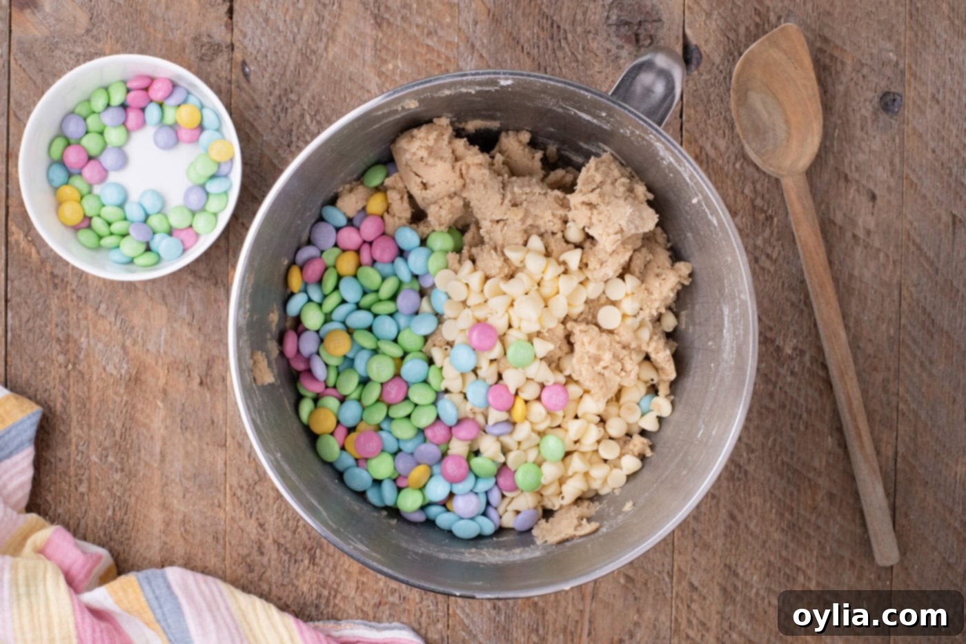 White chocolate chips and M&Ms being added to cookie dough in a bowl and folded in.