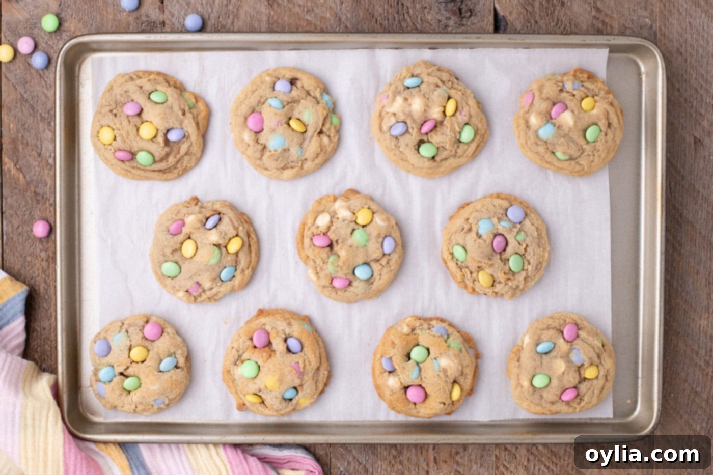 Overhead view of freshly baked Easter M&M cookies on a baking sheet, with extra M&Ms and chocolate chips being pressed into them.