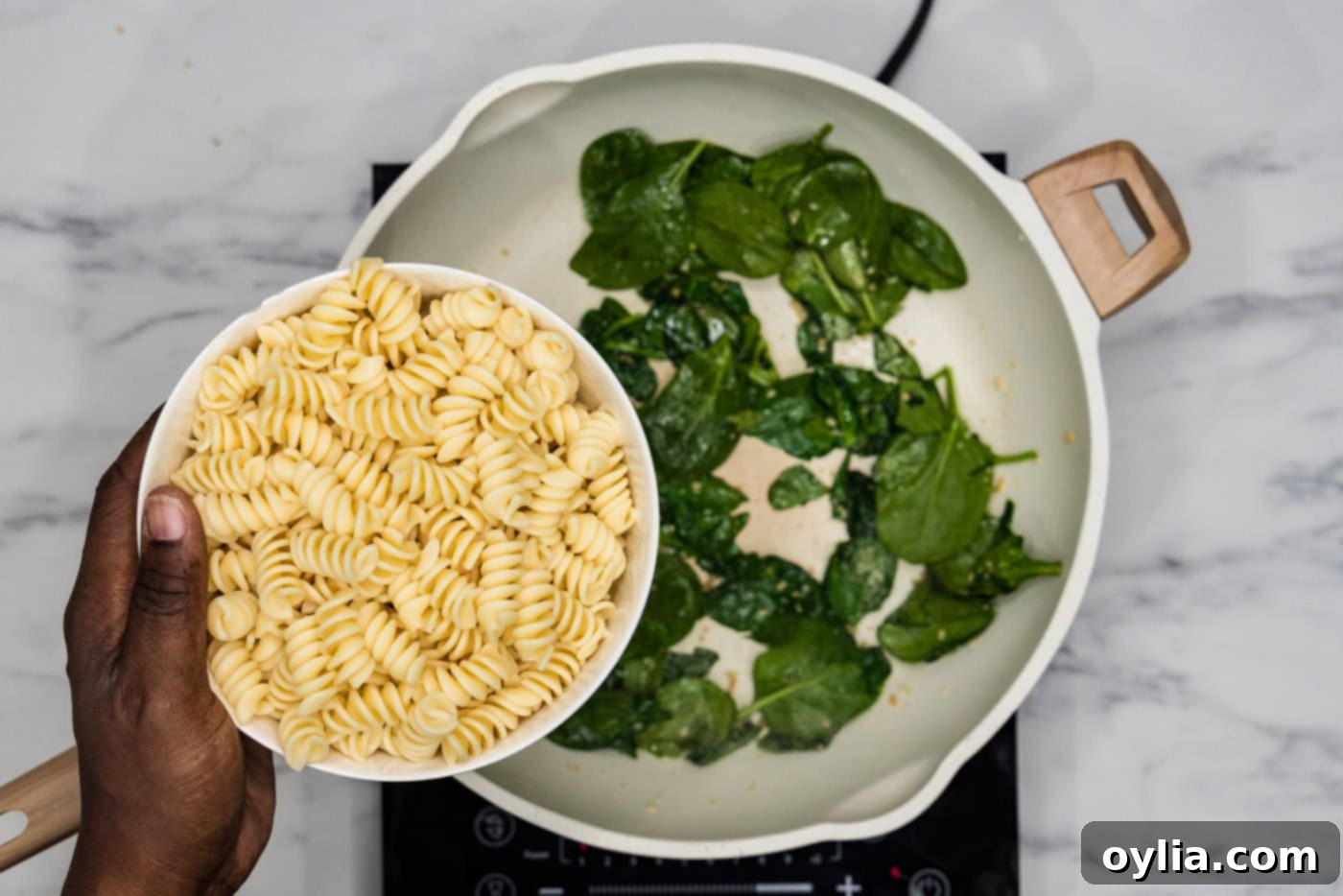 Adding cooked rotini pasta to the skillet with wilted spinach and garlic, ready to be tossed with pesto.