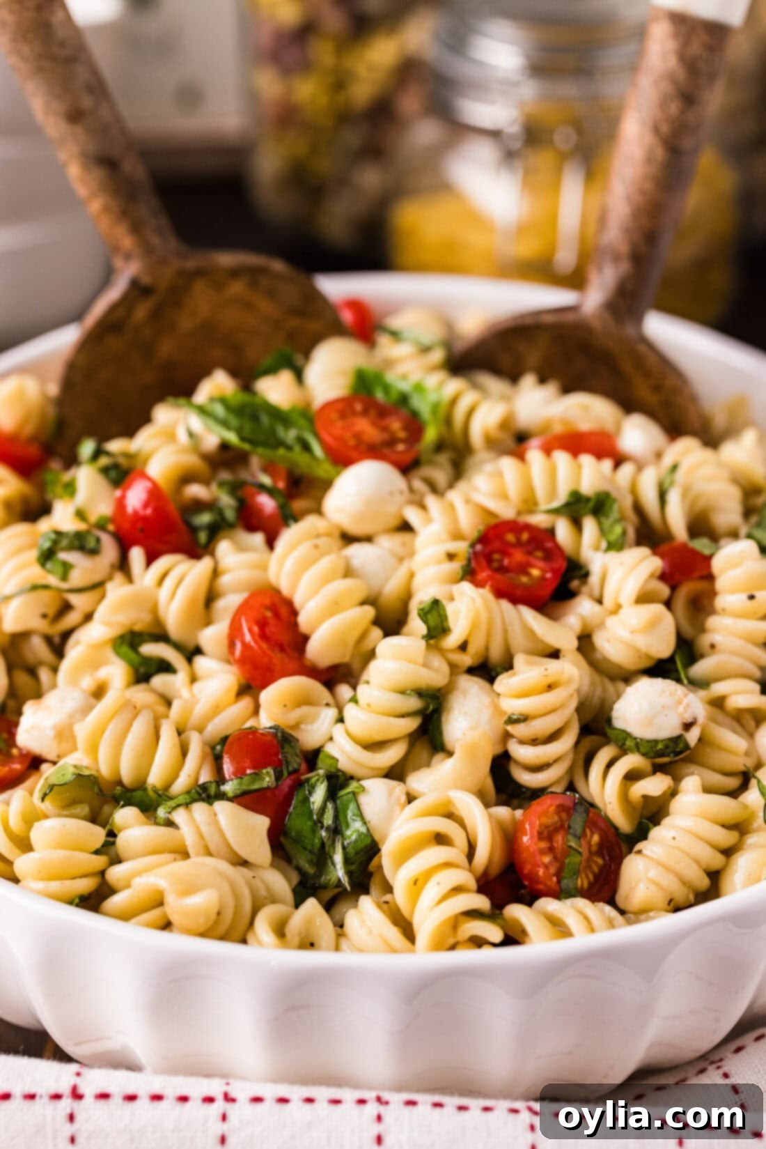 Two spoons elegantly placed in a large, inviting bowl of Caprese Pasta Salad, showcasing the colorful medley of cherry tomatoes, mozzarella pearls, and fresh basil.