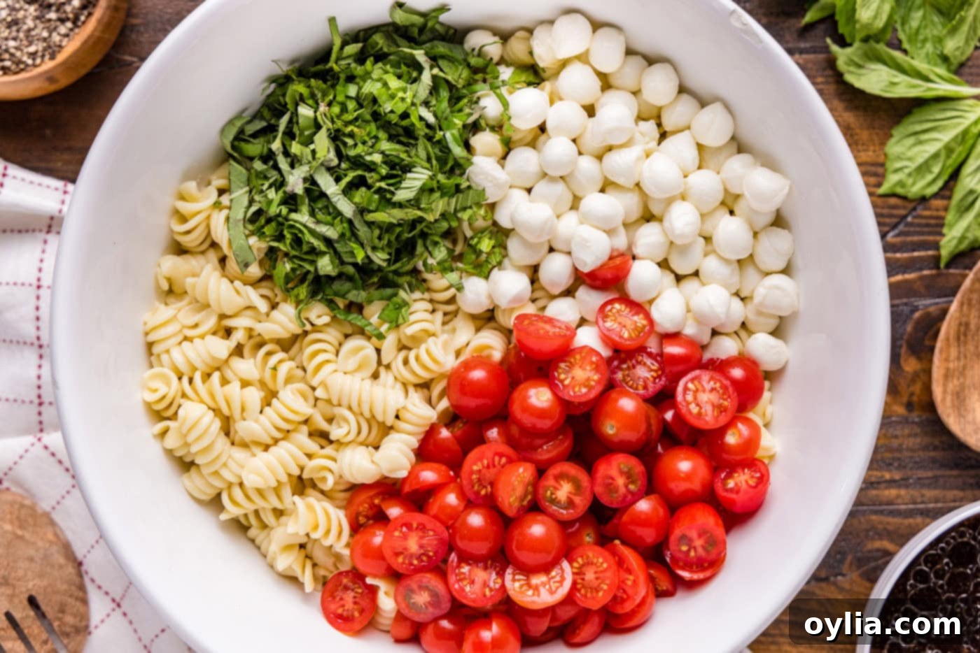 Chopped basil leaves, halved cherry tomatoes, and small mozzarella balls mixed with cooked rotini pasta in a large mixing bowl.