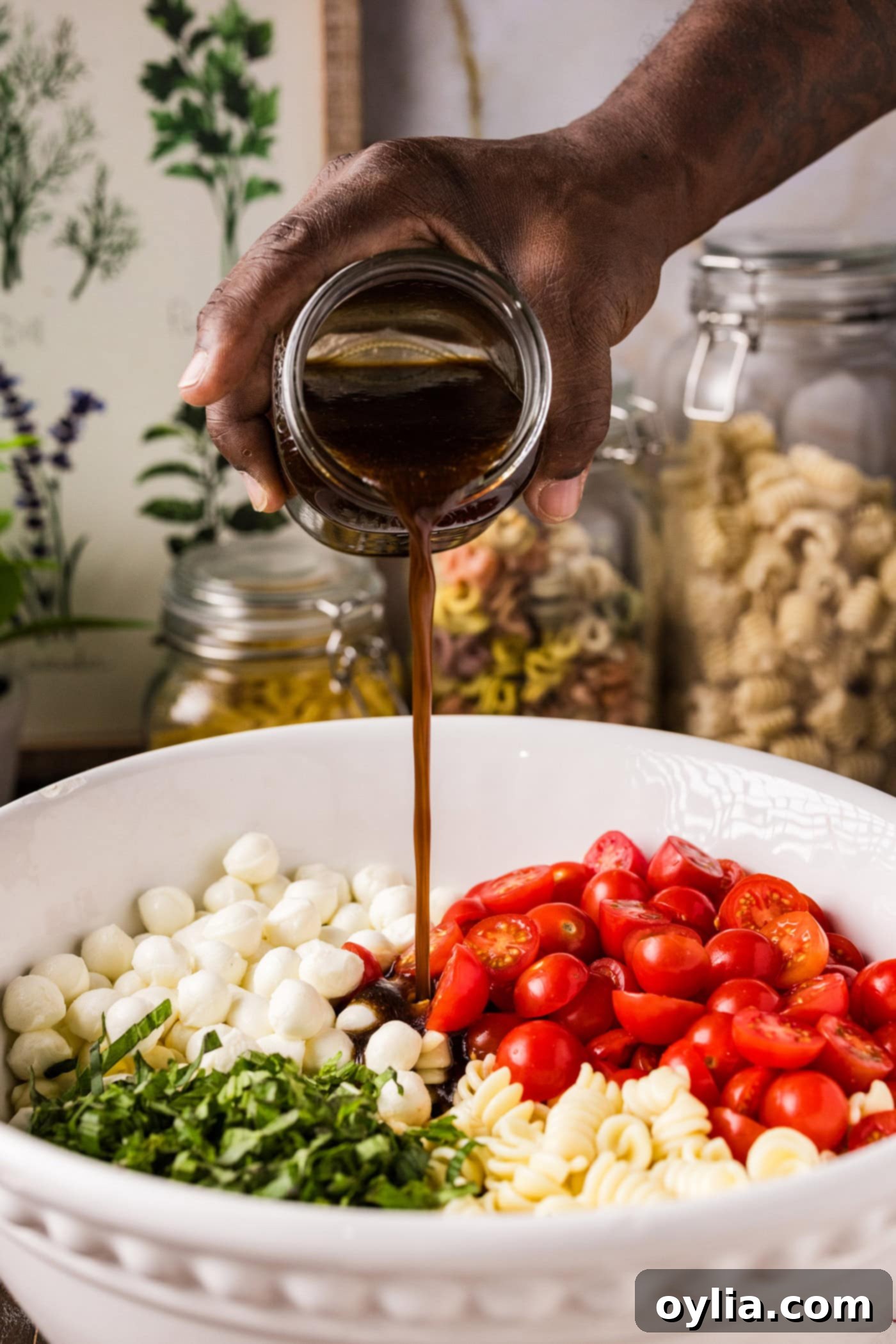 Balsamic vinaigrette being poured over the colorful Caprese Pasta Salad ingredients in a bowl, ready for tossing.