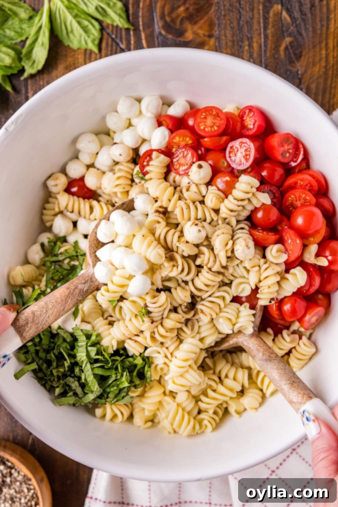 Caprese Pasta Salad being gently mixed in a large white serving bowl, showing the even distribution of ingredients and dressing.