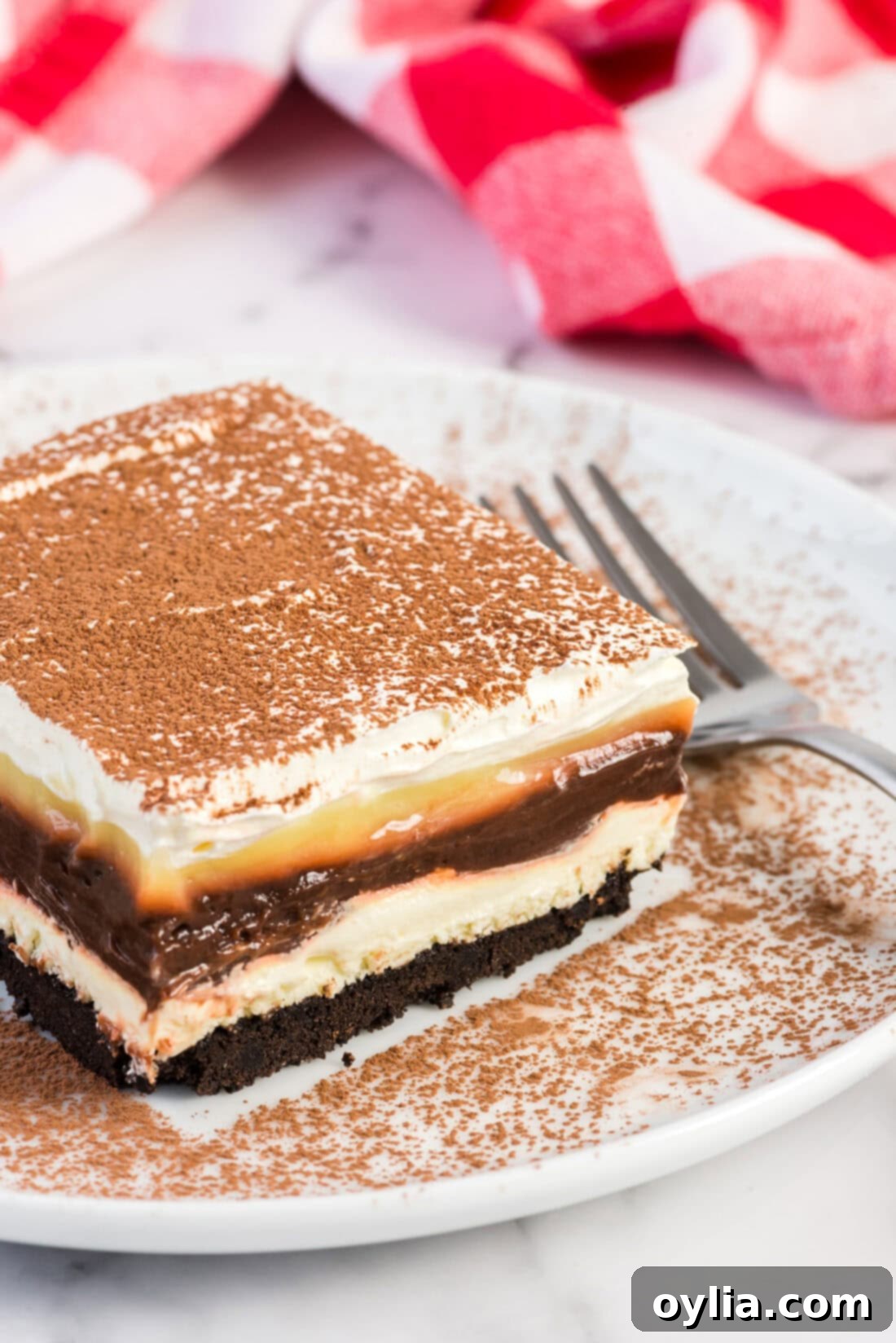 A perfectly sliced piece of Sex in a Pan dessert on a white plate, showcasing its distinct layers of crust, cream cheese, chocolate and vanilla pudding, and whipped topping, with a fork beside it.