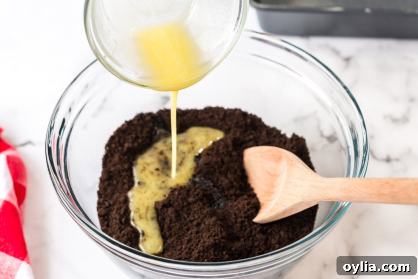 Melted butter being poured into a bowl of chocolate graham cracker crumbs, ready to be mixed for the crust.