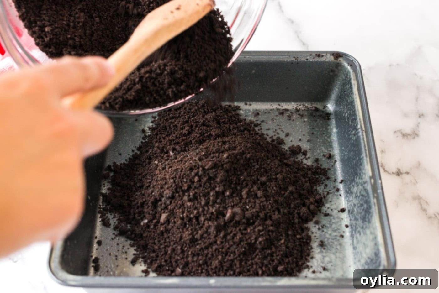 The chocolate graham cracker crumb mixture being pressed firmly into a 9x9 inch baking pan to form the dessert crust.