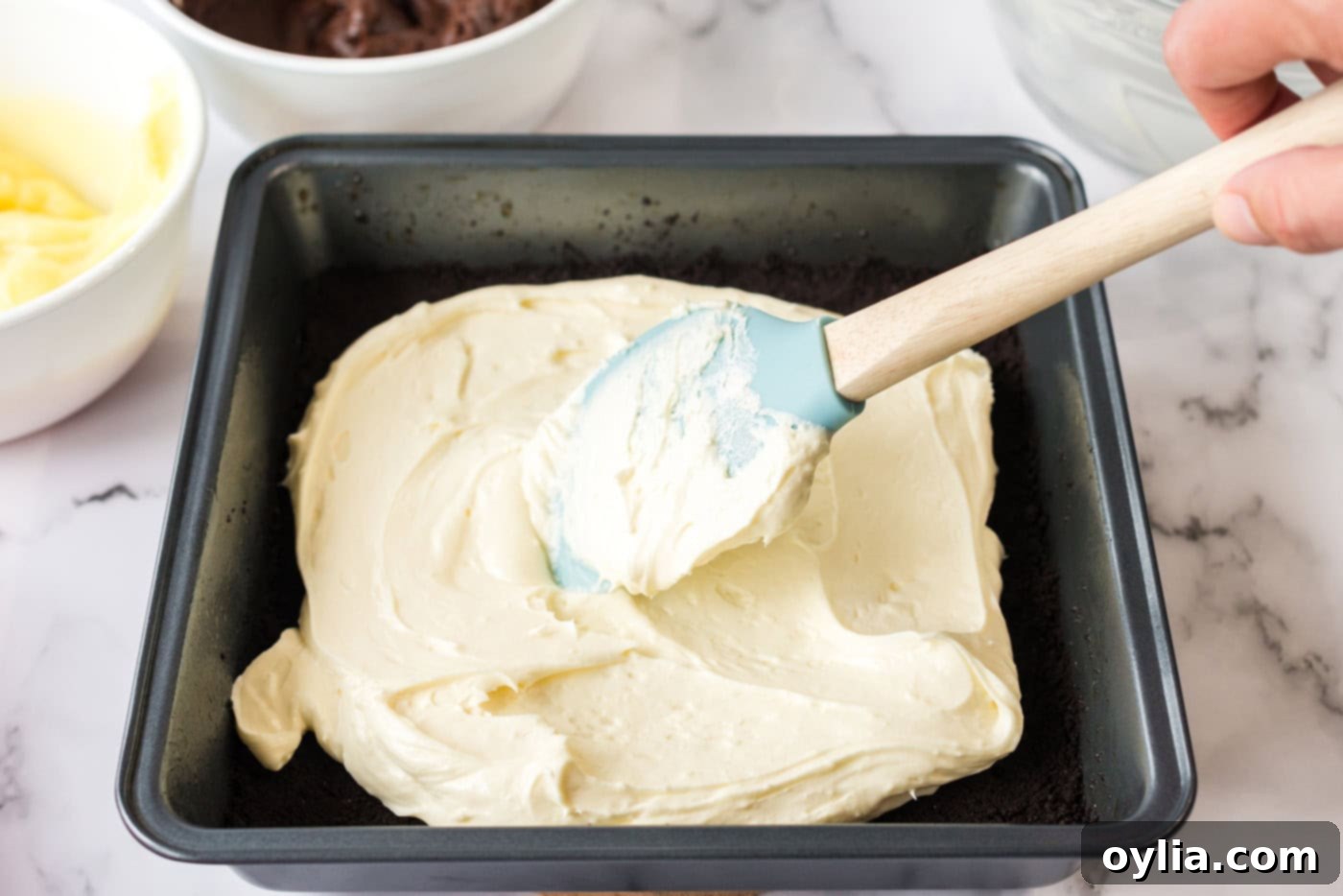 The creamy white cream cheese mixture being carefully spread evenly over the baked chocolate crust in a baking pan.
