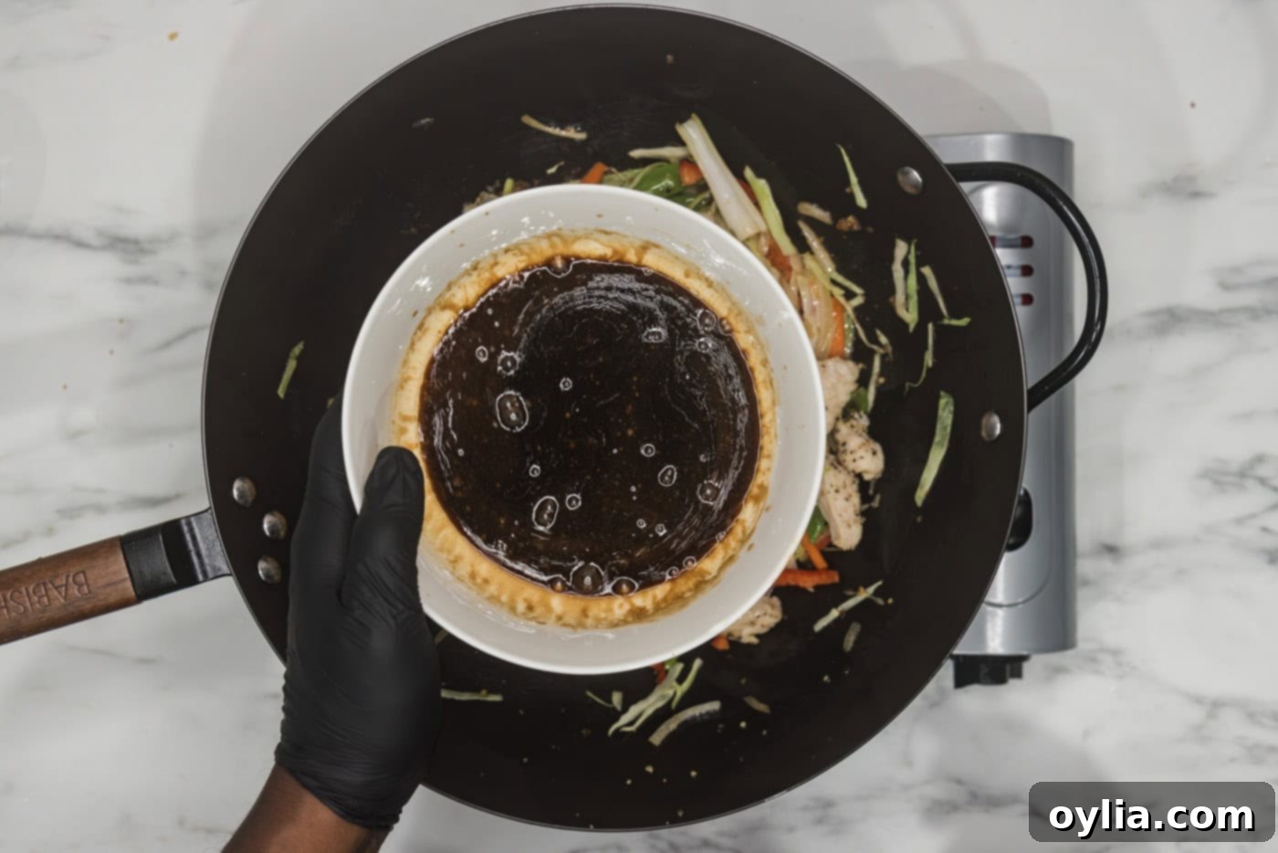 gloved hand holding lo mein sauce in a bowl over a wok