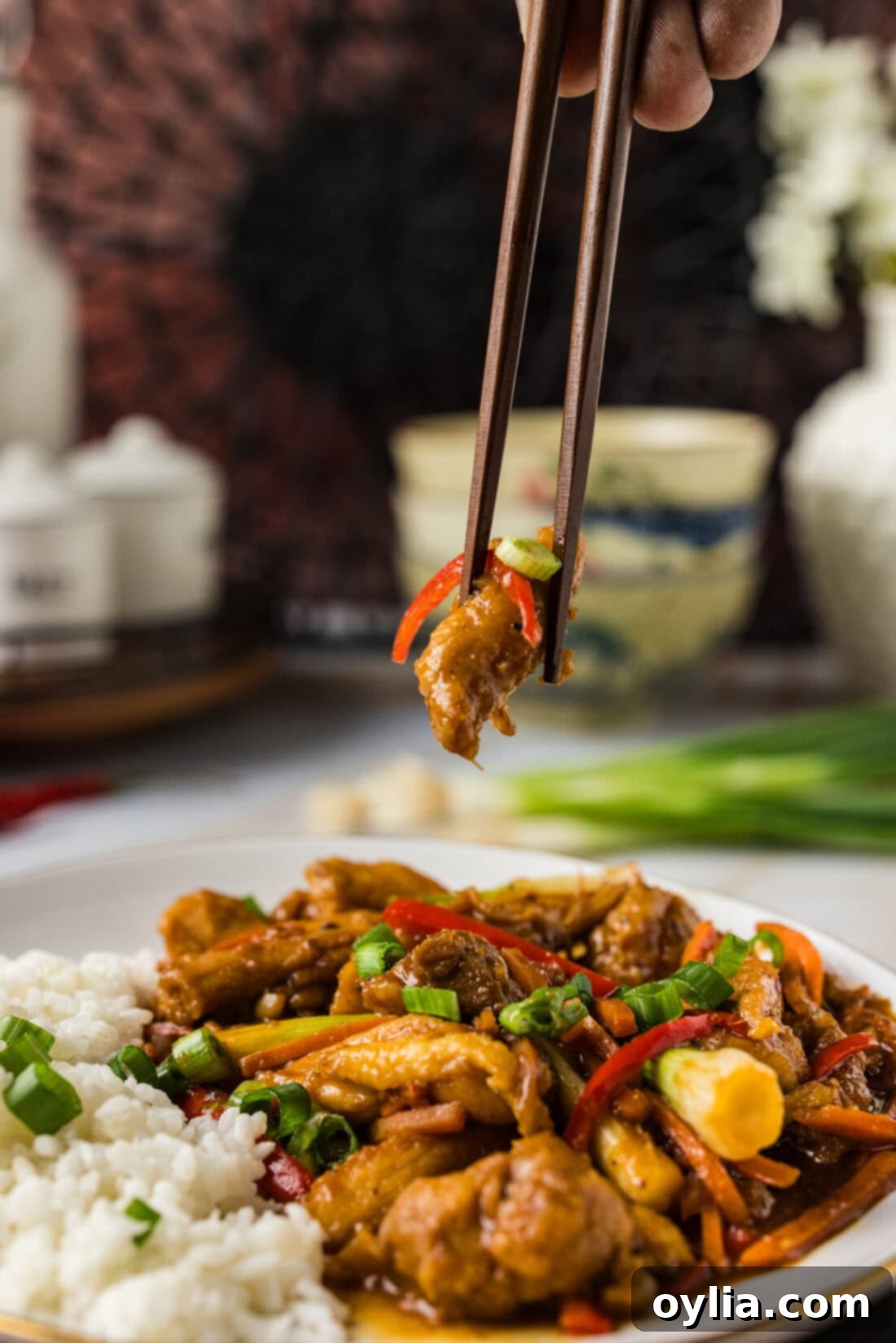Spicy Szechuan Chicken 10 Chopstick holding a piece of Szechuan Chicken over a plate of the dish, ready to be enjoyed.