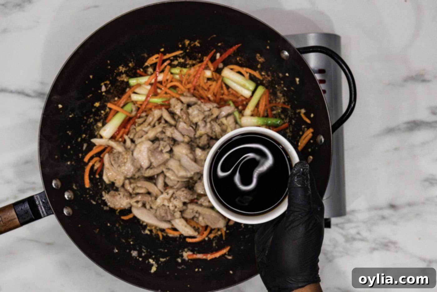 Gloved hand pouring soy sauce into a wok filled with Szechuan chicken and stir-fried vegetables.