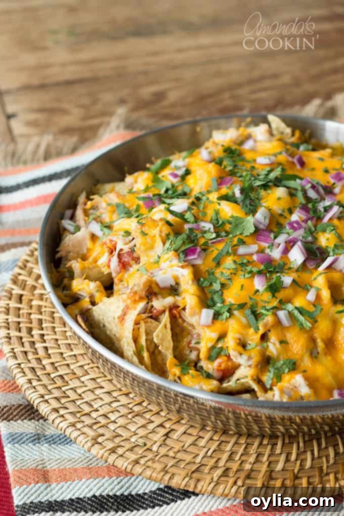 A stainless steel bowl of rotisserie chicken nachos resting on a wicker place mat, showing the generous toppings and melted cheese.