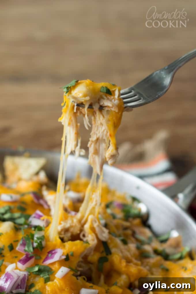 A photo of a fork pulling a piece of cheesy rotisserie chicken nachos from a pan, showing a luscious cheese pull.