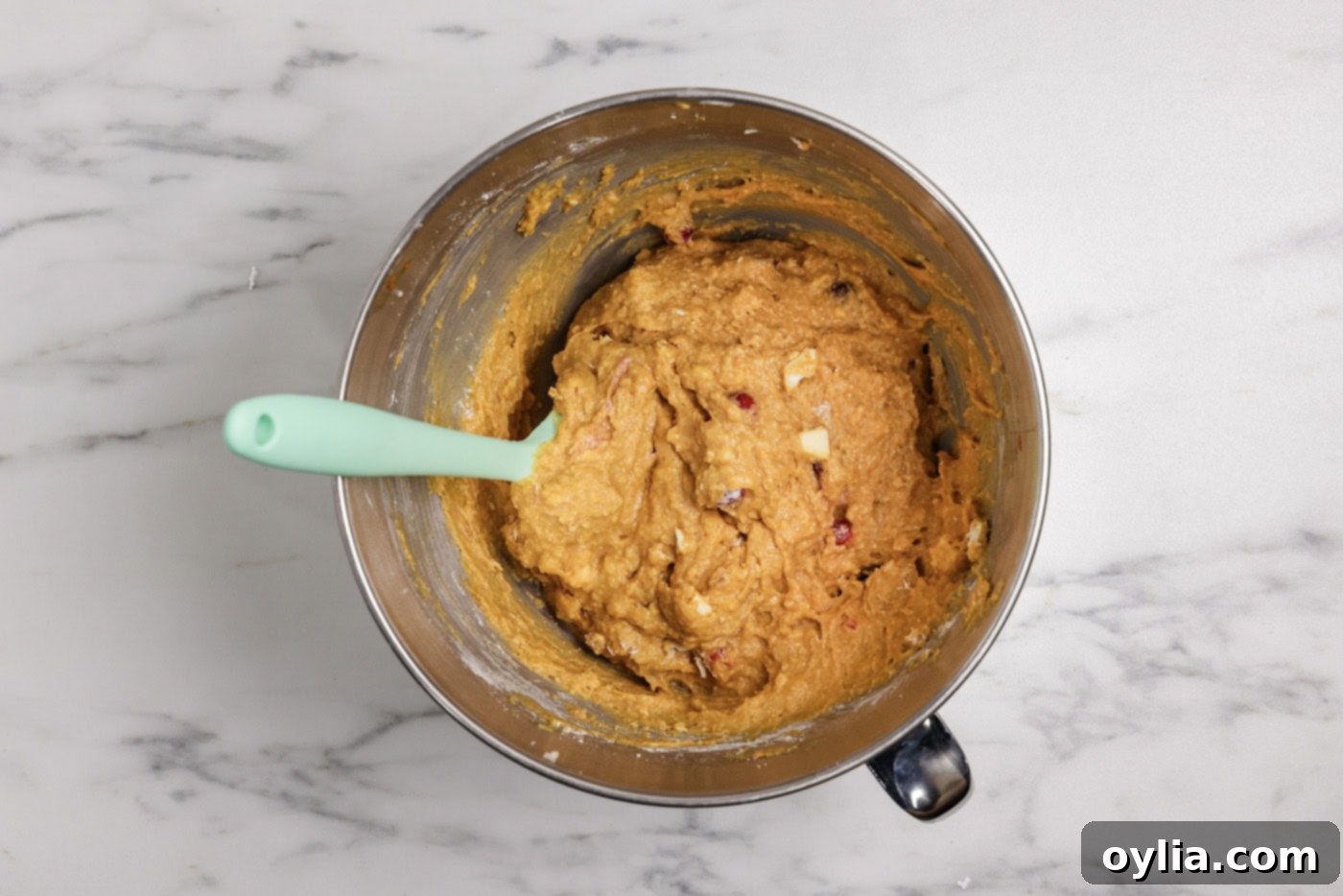 A rubber spatula gently folding cranberries and white chocolate into pumpkin muffin batter in a large bowl.