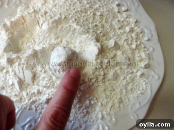 A raw chicken ball being rolled and coated thoroughly in the flour mixture on a white plate.