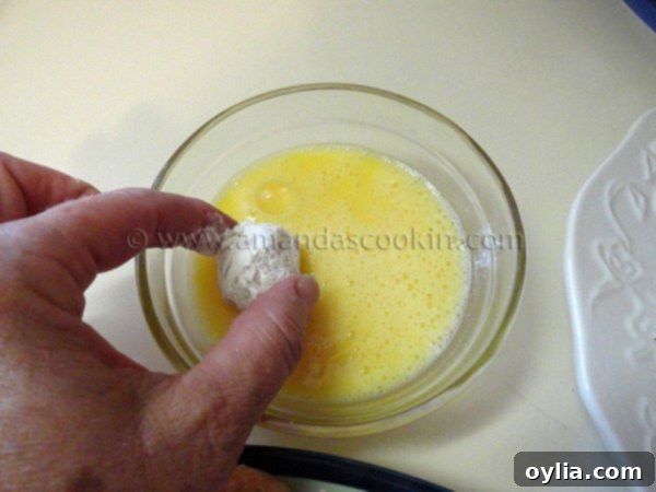 The floured chicken ball being dipped and coated evenly with whisked egg in a white bowl.