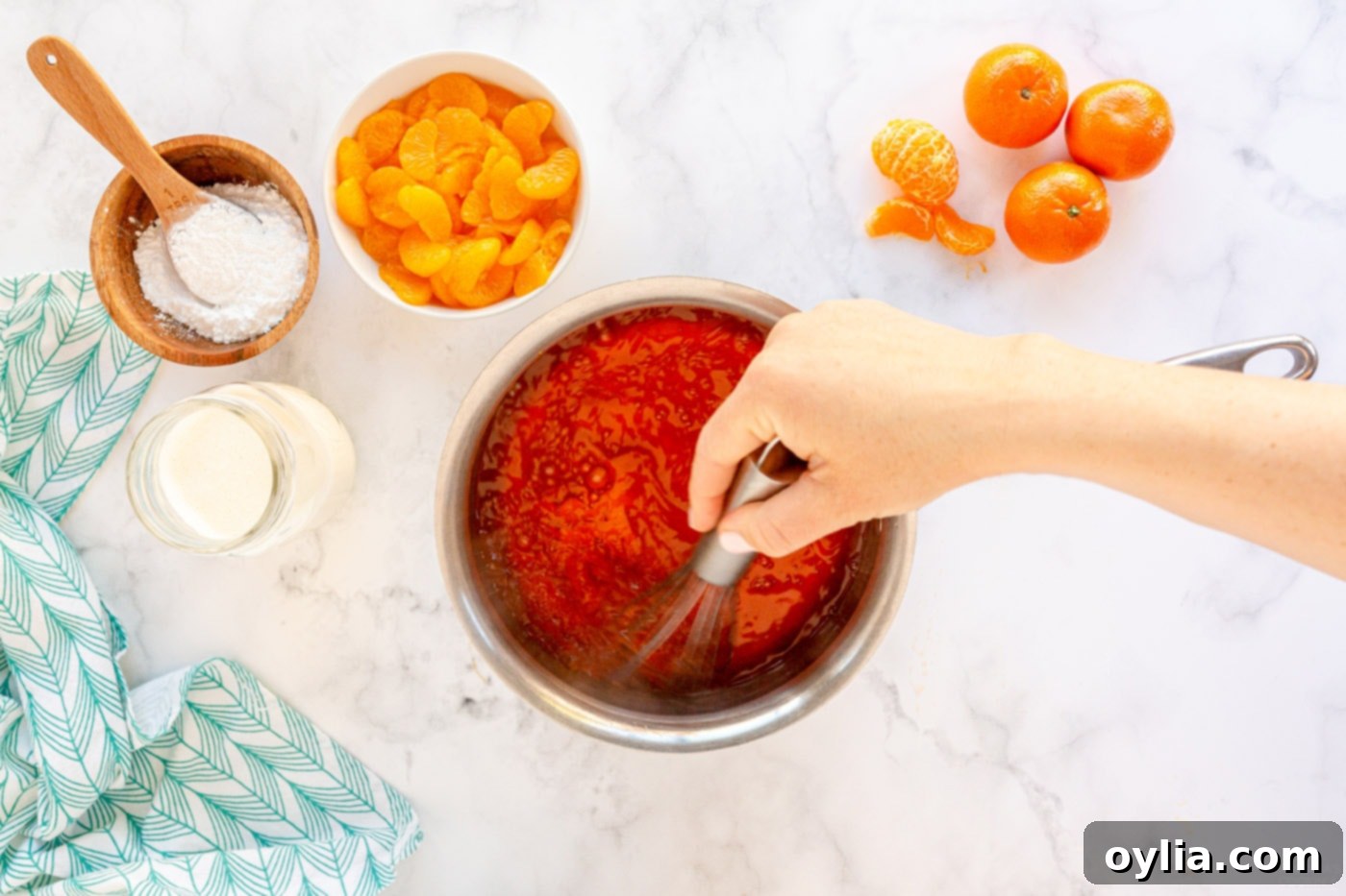 A saucepan on a stovetop with mandarin orange juice and water boiling, next to a packet of gelatin powder being stirred in until completely dissolved, indicating the first step of preparing the gelatin base.