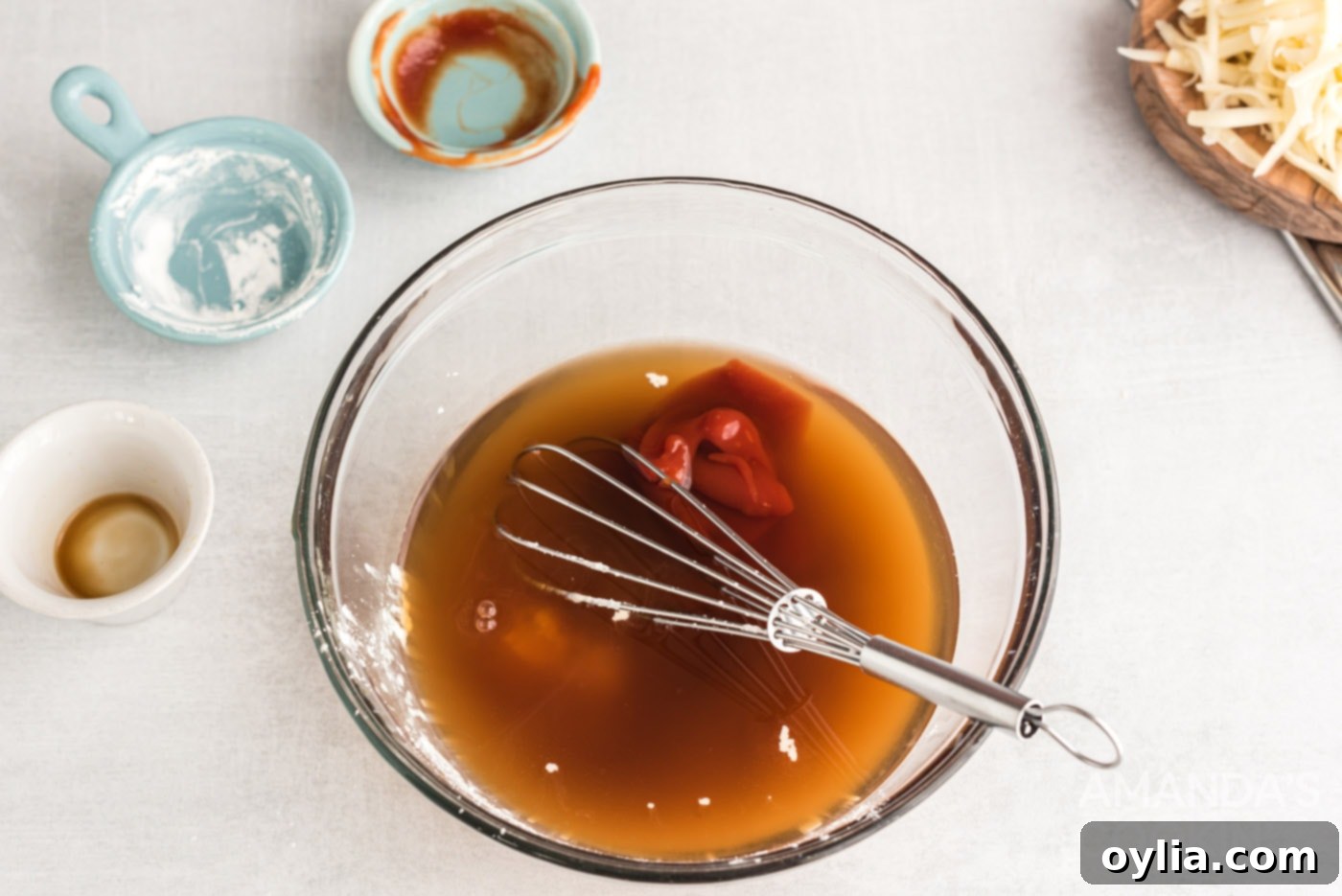 A bowl containing beef broth, Worcestershire sauce, and ketchup, with a whisk, showing the prepared sauce base