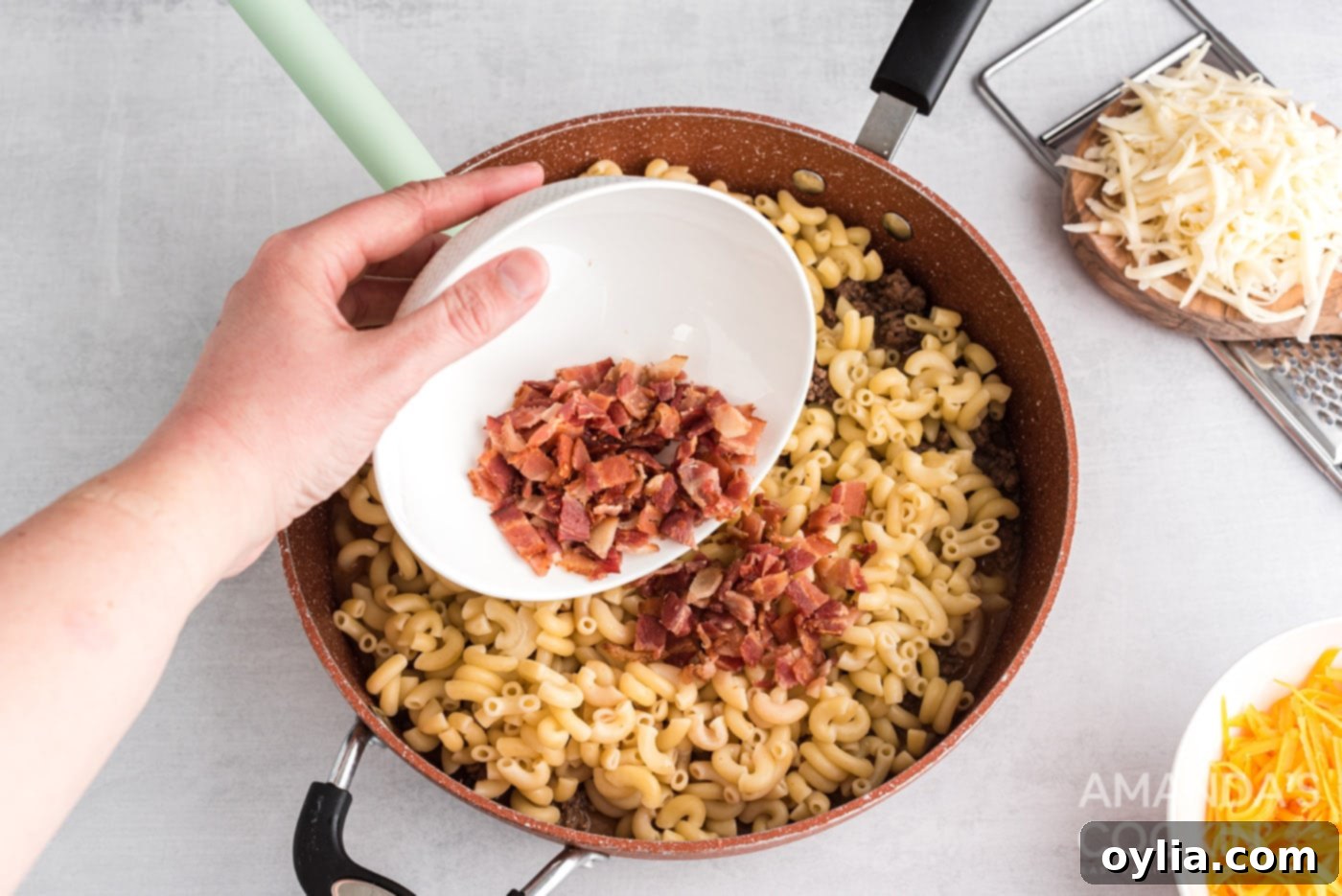 Adding crispy bacon bits to the skillet containing pasta and ground beef, ready for mixing
