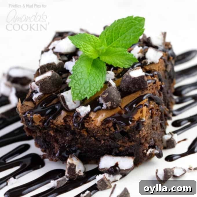 A close up photo of a peppermint patty brownie topped with drizzled chocolate sauce, chopped peppermint patties and a mint leaf.