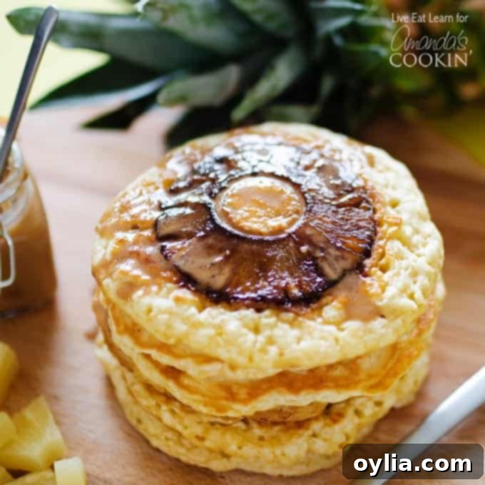 A close up photo of a stack of pineapple pancakes on a wooden cutting board, with warm coconut syrup drizzled over them and a pineapple ring visible.