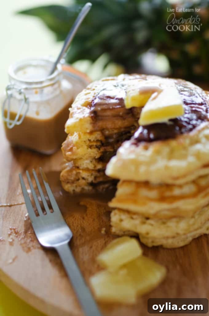 A close up vertical photo of a stack of pineapple pancakes cut into, revealing a pineapple ring inside, on a wooden cutting board. Drizzled with coconut syrup.