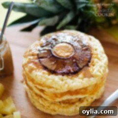 A close up photo of a stack of pineapple pancakes on a wooden cutting board, with warm coconut syrup drizzled over them and a pineapple ring visible.