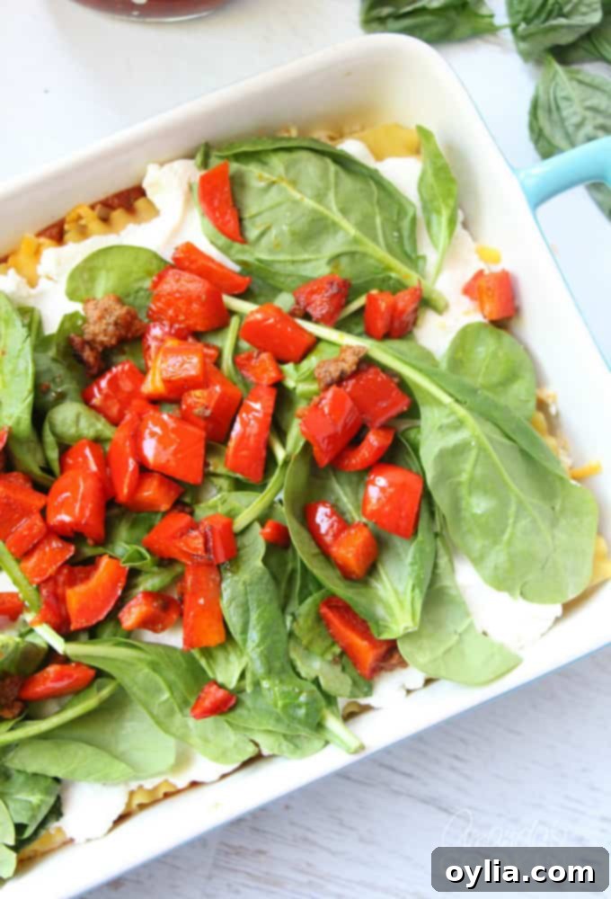 A photo showcasing a layer of ricotta cheese on lasagna noodles, followed by fresh spinach, and then bright chopped red peppers on top.