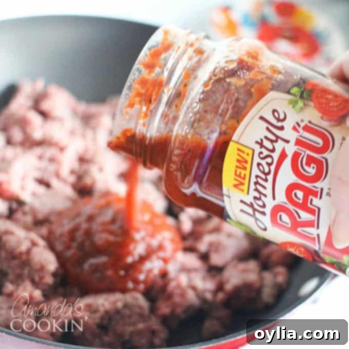A close up photo of Ragu Homestyle sauce being poured into a skillet with browned ground beef, mixing ingredients for the first layer.