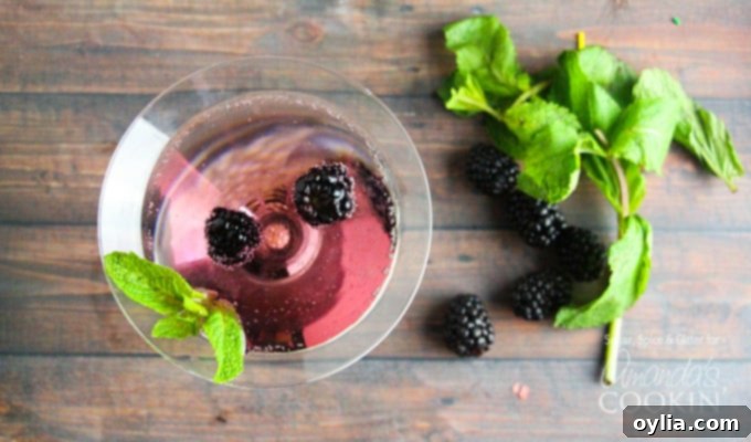 An overhead of a blackberry gin martini topped with fresh blackberries and mint leaves and some on the table next to the glass.