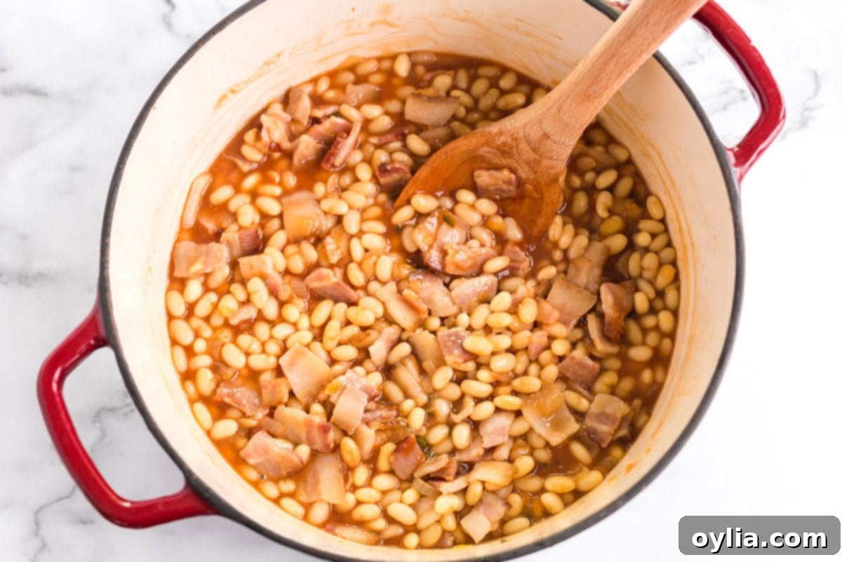 These homemade baked beans are baked in a delicious tangy sweet and savory sauce of brown sugar, maple syrup, ketchup, and apple cider vinegar. A close-up shot of a wooden spoon gently stirring the rich, dark sauce of homemade baked beans in a Dutch oven.